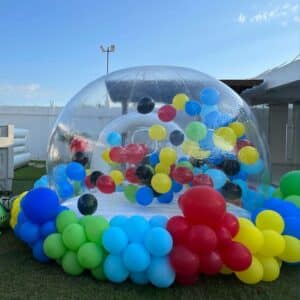 Burbuja inflable para jardín decorada con globos de colores llamativos debajo de una pérgola de madera.